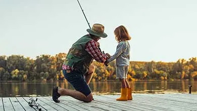 A man in a fishing vest and hat kneels on a wooden dock, assisting a young child in yellow boots with a fishing rod.