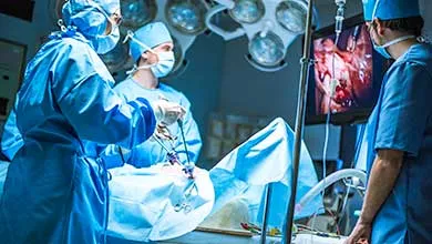Surgeons in blue scrubs and masks perform a laparoscopic surgery in an operating room, while one watches the procedure on a monitor.