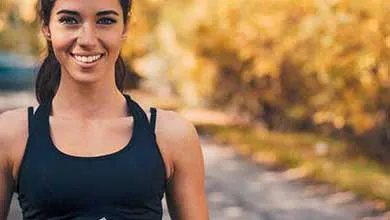 A young woman with dark hair pulled back, smiling, wears a black sports top outdoors against a backdrop of golden autumn leaves.