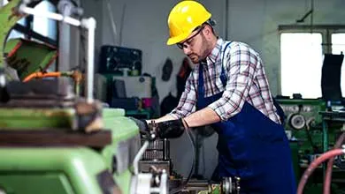 A man wearing a yellow hard hat, glasses, and a blue apron operates a large green machine in a workshop.