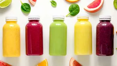 Five bottles of juice in yellow, red, green, and purple are lined up on a white background, surrounded by fresh fruit slices and spinach leaves.