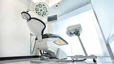 A modern dental operatory with a white reclining chair featuring black upholstery, positioned under a circular LED surgical light. An adjustable computer monitor is mounted above the chair, and a mobile dental unit on wheels is to the right. The room has white walls, large windows with bright natural light, and a clean, clinical appearance.
