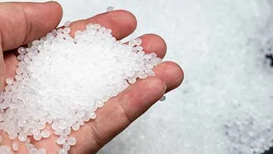 Hand holding translucent white plastic pellets with additional pellets scattered in the background.