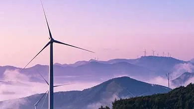 A large wind turbine stands in the foreground with mist-covered mountains behind it at twilight, while smaller turbines line the distant ridges under a soft pink and purple sky.